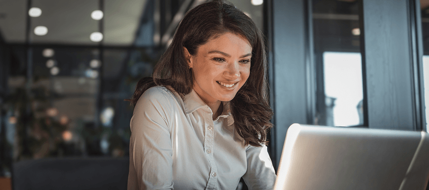 A professionally dressed woman smiles at her laptop screen.