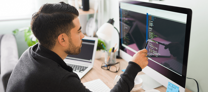 A web developer working at a desk, reviewing code on a large monitor with a laptop and coffee nearby.