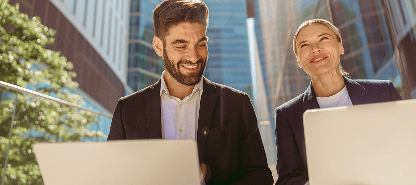 Two smiling real estate agents holding laptops while walking down the street of a business district.