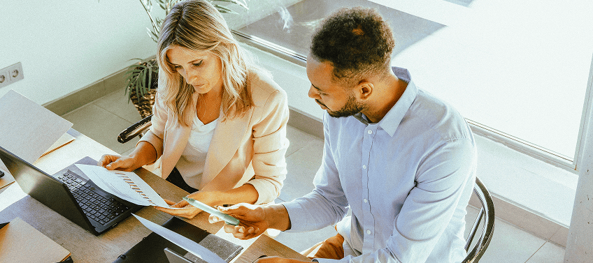 A male and a female professional in a conference room, working on their laptops and comparing notes.